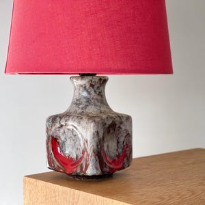 Puede incluir: Una lámpara de mesa vintage con una pantalla roja. La base de la lámpara tiene forma cuadrada con un acabado moteado gris y marrón, acentuado con detalles rojos. La lámpara está colocada sobre una superficie de madera.