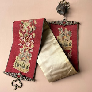 May include: A vintage red tapestry with a castle and bird design, framed with ornate metalwork. The tapestry is paired with a gold satin fabric. A small bell hangs above the tapestry.