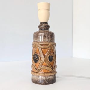 Puede incluir: Lámpara de mesa de cerámica vintage con casquillo beige. La lámpara tiene un cuerpo moteado marrón y canela con una banda naranja con un patrón decorativo. Un cable eléctrico blanco se extiende desde la base.