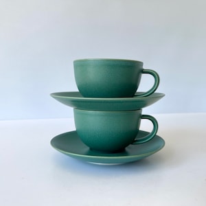 May include: Two green ceramic teacups and saucers stacked on top of each other. The cups have a rounded shape and a slightly textured surface.