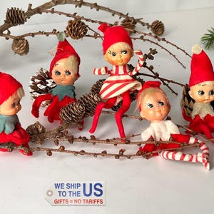 Peut inclure: Cinq figurines d'elfes de Noël vintage avec des chapeaux et des tenues rouges, posées sur une branche avec des pommes de pin. Les elfes ont des visages peints et sont dans diverses positions. Une brindille de conifère est en arrière-plan.