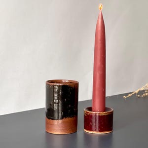 May include: A ceramic candle holder with a lit burgundy taper candle. Beside it is a matching ceramic cup. The candle holder and cup have a dark brown and black glaze over a reddish-brown base. Home decor.