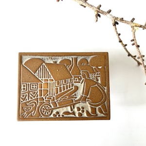 Puede incluir: Un azulejo de cerámica marrón con un diseño blanco de una casa, una persona empujando una carretilla y árboles. El azulejo tiene un aspecto rústico y vintage.