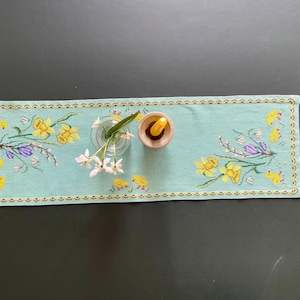 Puede incluir: Camino de mesa rectangular bordado en azul claro con narcisos amarillos, flores moradas y pollitos amarillos. Un pequeño jarrón de cristal con flores blancas y un cuenco de madera con una vela encima. El camino tiene un borde dorado.