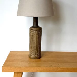 Pode incluir: Um candeeiro de mesa com uma base cilíndrica alta de cerâmica com um padrão castanho texturado em forma de grelha e um pescoço liso e cónico. O candeeiro é rematado com um abajur de tambor em tecido bege claro, simples e sólido.