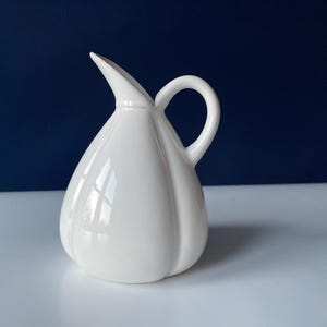 May include: A white ceramic pitcher with a curved handle and a spout. The pitcher has a bulbous body with vertical ridges and a glossy finish. The background is a dark blue.