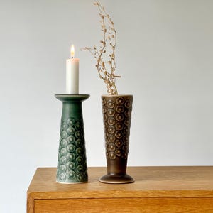 Puede incluir: Un portavelas de cerámica verde con una vela blanca encendida y un jarrón marrón con un estampado floral. El portavelas y el jarrón están sobre una superficie de madera.