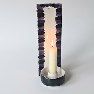 May include: A white ceramic candle holder with a green base and a black and purple patterned border. A white candle is lit inside the holder.