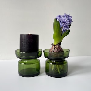Può includere: Due portacandele in vetro verde, uno con una candela nera accesa e l'altro con un fiore di giacinto viola. I portacandele hanno una base rotonda e una parte superiore circolare.