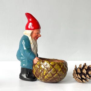 May include: A ceramic garden gnome figurine with a red hat and blue coat, holding a pineapple-shaped planter. A pine cone rests nearby. The gnome is a whimsical decorative item.