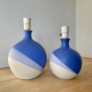 May include: Two ceramic table lamps with a blue and white color scheme. The lamps have a rounded base and a diagonal color block design. The lamps are of different sizes, with a white lamp holder at the top.