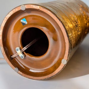 May include: Close-up of the bottom of a brown ceramic lamp base with a gold metallic glaze. The base has a round opening for the cord and a label that reads "BERNDORFER".