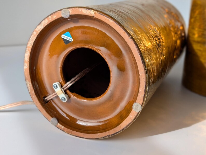 May include: Close-up of the bottom of a brown ceramic lamp base with a gold metallic glaze. The base has a round opening for the cord and a label that reads "BERNDORFER".