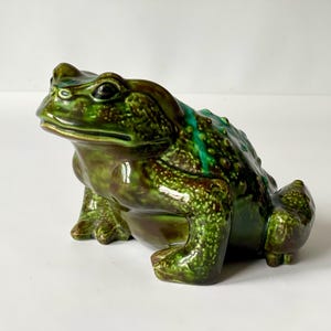 Puede incluir: Figura de rana de cerámica en tonos verdes y marrones. La rana tiene piel texturizada, ojos detallados y está en posición sentada. La figura es un artículo decorativo, adecuado para la decoración del hogar.