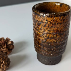Peut inclure: Une petite tasse en céramique brune avec un glaçage texturé et moucheté. La tasse a un fond arrondi et un bord légèrement évasé.