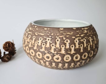 Op de afbeelding: Een bruine keramische kom met een wit interieur en een herhalend patroon van beige cirkels en abstracte vormen.