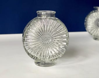 May include: Clear glass decorative vase with a circular shape and a textured, sunburst-like design. The vase has a small, round base and a narrow neck. The background is a deep blue, and the vase is sitting on a white surface.