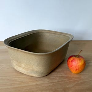 May include: A rectangular ceramic bowl with rounded corners and a matte, light beige finish. The bowl has a wide, open top and a smooth, uniform texture. A single red and yellow apple is placed next to the bowl for a visual size comparison.