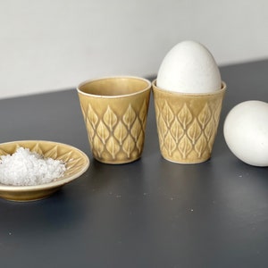 Op de afbeelding: Twee groene keramische eierdopjes met een bladmotief, een met een wit ei erin, en een klein groen schoteltje met zout.