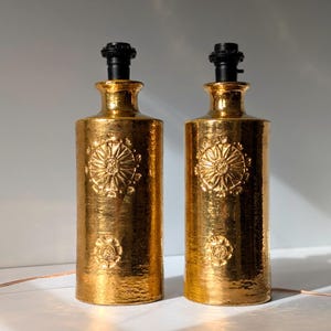 May include: Two gold ceramic table lamps with black lampshades. The lamps have a textured surface and a floral design.