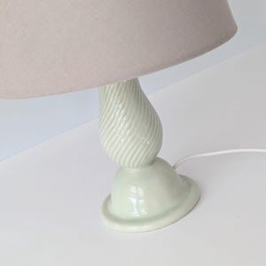 May include: A small, decorative table lamp with a light beige lampshade. The base is a pale green, ceramic design with a spiral pattern. The lamp is plugged into a white electrical cord.