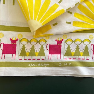 May include: White fabric with a repeating pattern of yellow sunbursts and a border featuring pink cows and figures holding hands. The words "spec. design. D.M.F." are printed on the border. The fabric is folded.