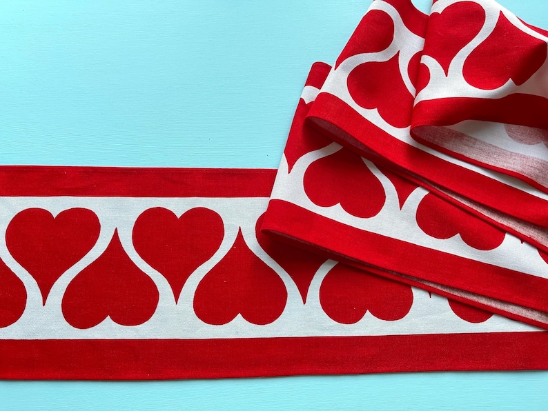 May include: Red and white fabric trim with a repeating pattern of white hearts on a red background. The trim is folded in thirds, showing the pattern from different angles.