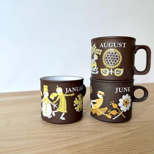 May include: Three brown ceramic mugs with white interiors are stacked on a wooden surface. The mugs are decorated with yellow and white illustrations and the months "January", "June", and "August".