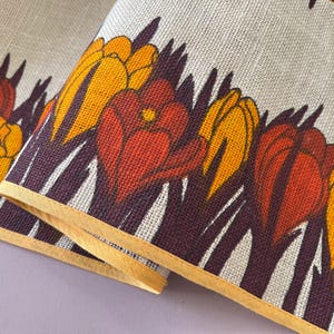 May include: A white linen fabric with a brown and orange floral pattern. The fabric has a yellow border and features a repeating pattern of stylised flowers.
