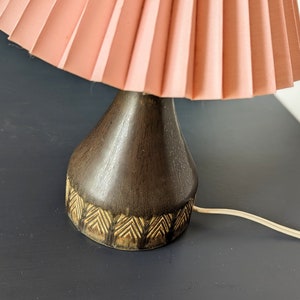 Puede incluir: Una lámpara de mesa vintage con una pantalla rosa plisada y una base de madera oscura con un patrón geométrico.