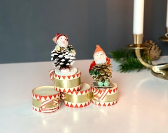 May include: Vintage Christmas decorations with gnome figures on pinecone bodies. The gnomes sit on small, round containers with red and gold striped ribbon. A festive holiday scene with a candle holder and greenery.