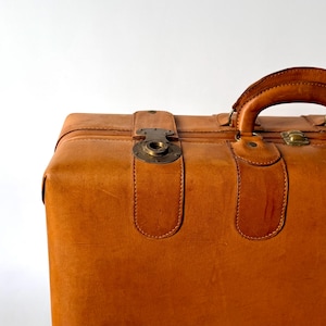 May include: A tan leather suitcase featuring a sturdy arched handle and brass-toned metal hardware. The exterior displays visible stitching along the reinforced leather straps and a circular metal latch mechanism centred on the top panel.