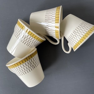 Puede incluir: Conjunto de cuatro tazas de café de cerámica blanca con un patrón geométrico amarillo y marrón. Las tazas están apiladas unas sobre otras.