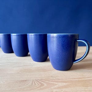 May include: Four cobalt blue ceramic mugs with a speckled texture. The mugs have a rounded shape and a handle. The mugs are arranged in a row on a light wood surface, with a dark blue background.