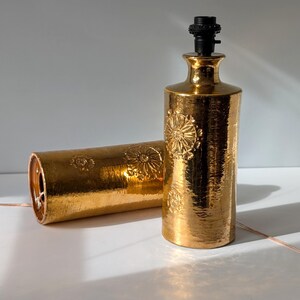 May include: Two gold ceramic table lamps with a floral embossed design. The lamps have a cylindrical shape and a simple design.