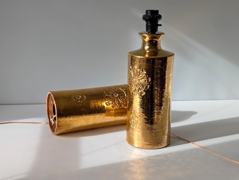 May include: Two gold ceramic table lamps with a floral embossed design. The lamps have a cylindrical shape and a simple design.
