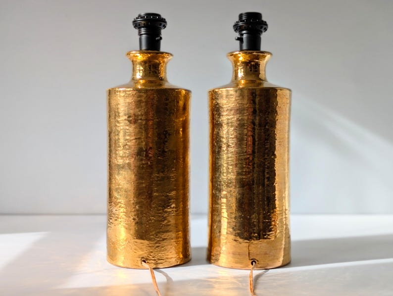 May include: A pair of gold ceramic table lamps with black lampshades. The lamps have a hammered texture.