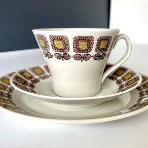 May include: A white teacup and saucer set with a brown and gold geometric pattern. The cup has a wide, rounded shape and a handle. The saucer has a matching pattern.