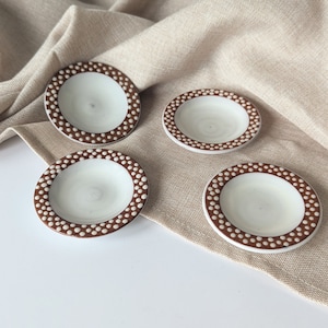 May include: Four small, round ceramic dishes with a white interior and a brown dotted border. The dishes are arranged on a beige linen surface, creating a simple, elegant aesthetic. These dishes are ideal for serving small portions of food or as decorative accents.
