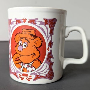 Könnte beinhalten: Weiße Keramiktasse mit einem roten, orangen und violetten Design. Die Tasse zeigt ein Cartoon-Bild eines Bären, der einen Hut und ein gepunktetes Hemd trägt.