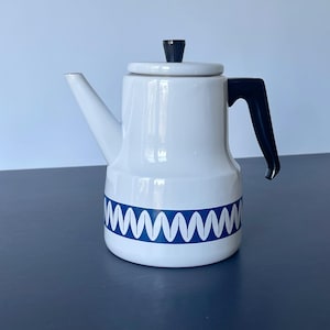 Puede incluir: Tetera de esmalte blanco con asa y pomo negros. La tetera tiene un patrón de zigzag azul y blanco alrededor de la base. El pico es blanco e inclinado.