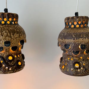 Peut inclure: Deux lampes suspendues en céramique émaillée brune avec une surface texturée et tachetée. Chaque lampe présente une série de découpes circulaires qui laissent passer une lumière chaude, créant un effet lumineux sur le corps brun foncé.
