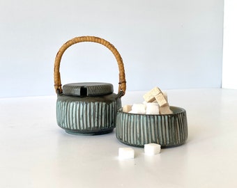 May include: A blue and white striped ceramic sugar bowl and creamer set with a woven handle on the creamer. The sugar bowl is filled with white sugar cubes.