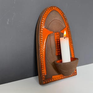 May include: A ceramic wall sconce with a lit white candle. The sconce has a brown and orange design with a curved arch shape. The candle sits in a small, round holder.