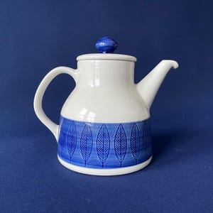 Könnte beinhalten: Weiße Keramik-Teekanne mit blauem Blättermuster am Boden und blauem Knauf auf dem Deckel. Die Teekanne hat einen gebogenen Griff und einen Ausguss. Der Hintergrund ist einfarbig dunkelblau.