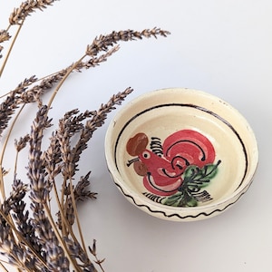 Può includere: Una piccola ciotola in ceramica bianca con un disegno dipinto a mano di un gallo rosso. La ciotola ha un bordo marrone e un bordo bianco e nero.