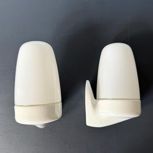 May include: Two off-white ceramic wall sconces. Each sconce has a rounded top and a rectangular base. One sconce has a curved arm extending from the base. The sconces are set against a dark background.