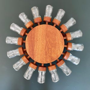 Puede incluir: Un especiero de madera con un diseño circular, con un disco central de madera y botes de especias de vidrio transparente dispuestos alrededor. Los botes tienen tapas de madera y están espaciados uniformemente, creando un efecto de sol.
