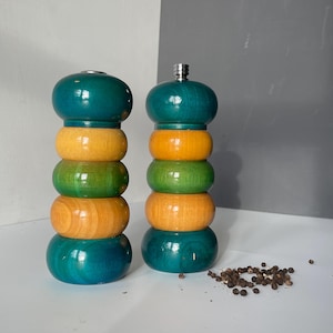 May include: A set of two wooden salt and pepper shakers with a teal, yellow and green layered design. The shakers are on a white surface with black peppercorns scattered around them.