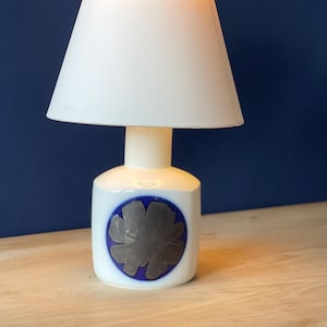 May include: A white table lamp with a conical lampshade. The base is white with a blue circle and a silver floral design. The lamp is on a wooden surface against a dark blue background.